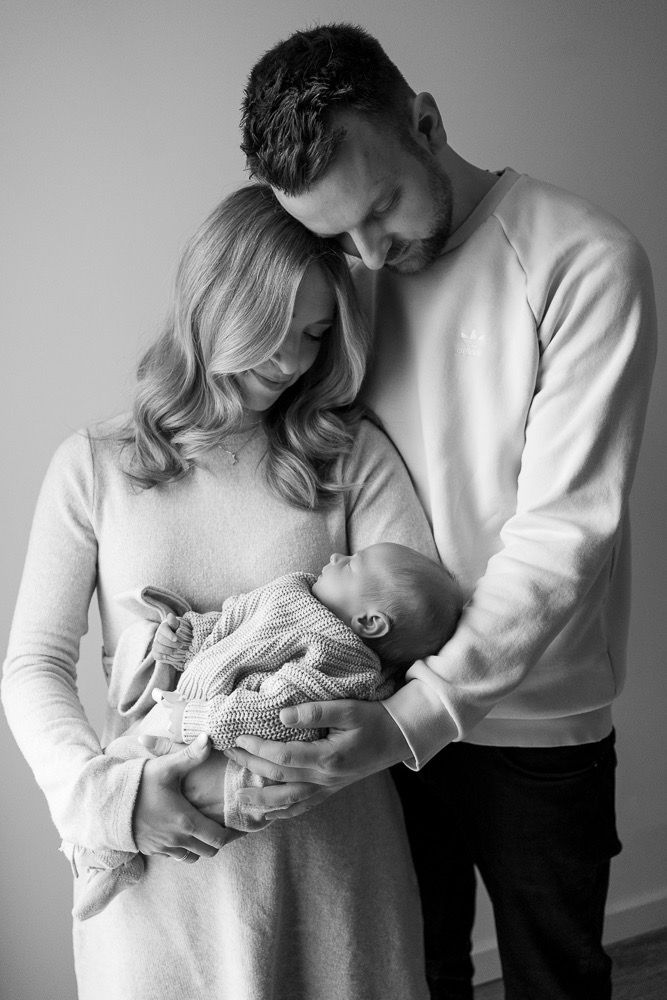 Black and white portrait of parents tenderly holding newborn wrapped in knit sweater.