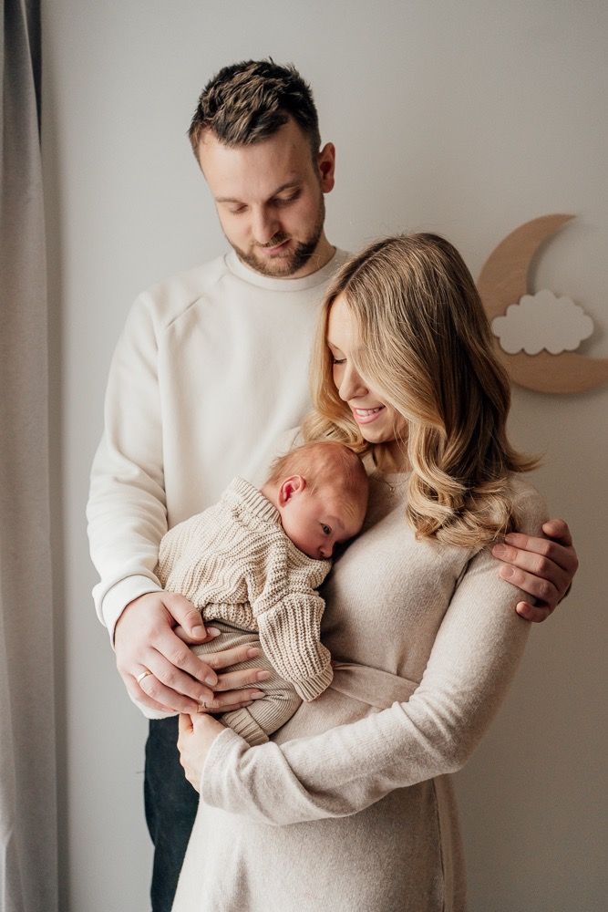 A couple in cream sweaters holds their newborn baby near a window in a nursery with wooden moon decor.
