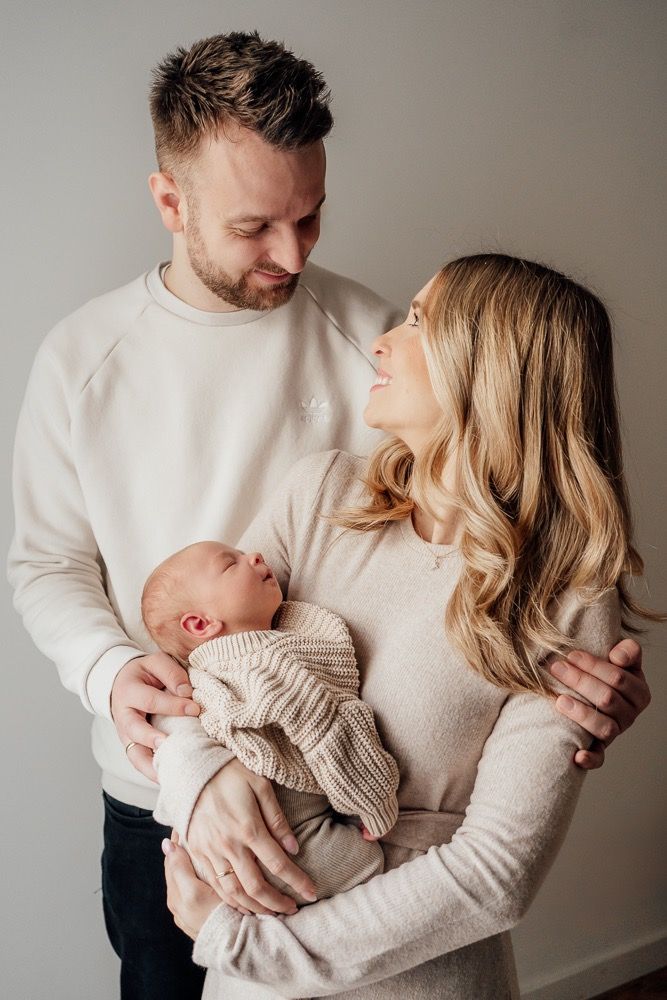 A family in cream colored sweaters share tender moments with their newborn baby in a minimalist studio setting.