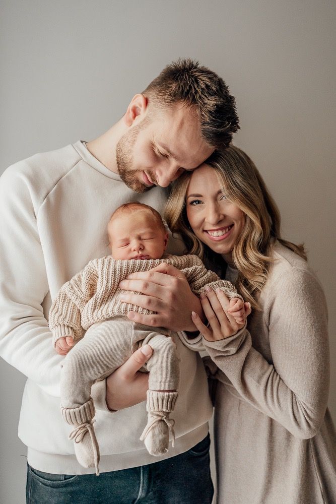 A tender family moment with parents in neutral sweaters cradling their newborn baby against a gray background.