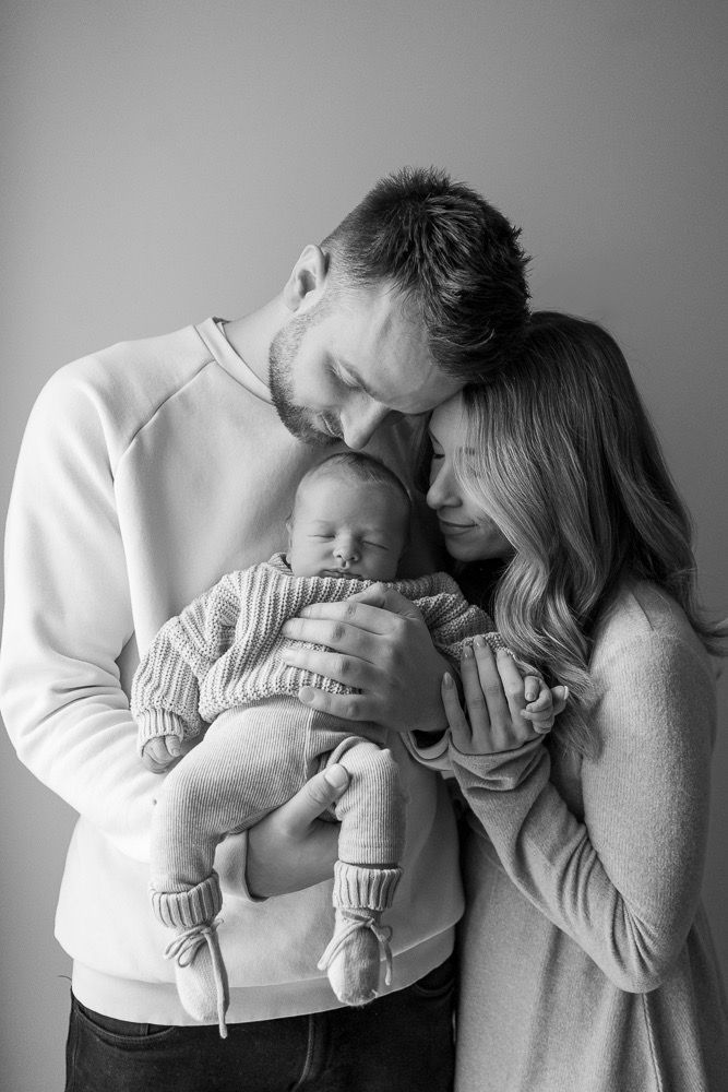 Black and white photo series showing tender moments between parents and their newborn baby in matching white outfits.