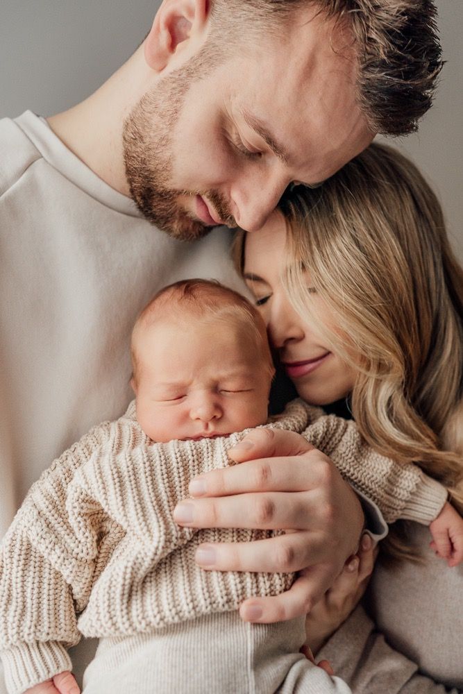 Intimate family portrait showing parents in soft sweaters embracing their sleeping newborn baby.