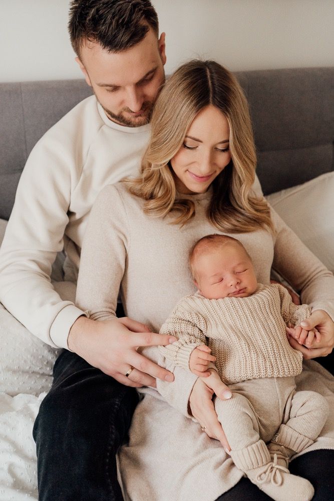 Parents in matching cream sweaters cuddle with their newborn baby on a gray couch in a tender family moment.