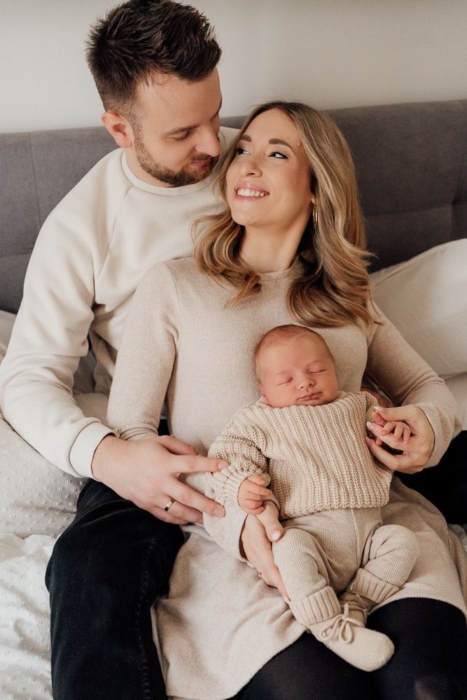 Family relaxing on gray couch in coordinated neutral outfits with sleeping newborn baby.
