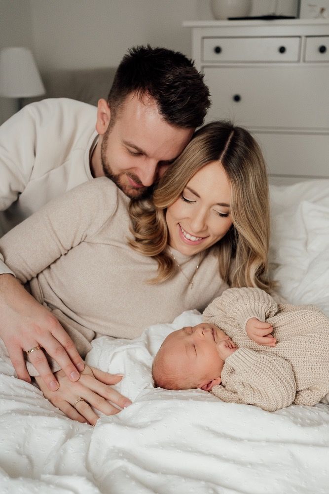 Parents lying in white bedding admiring their newborn baby in cozy neutral-toned bedroom setting.
