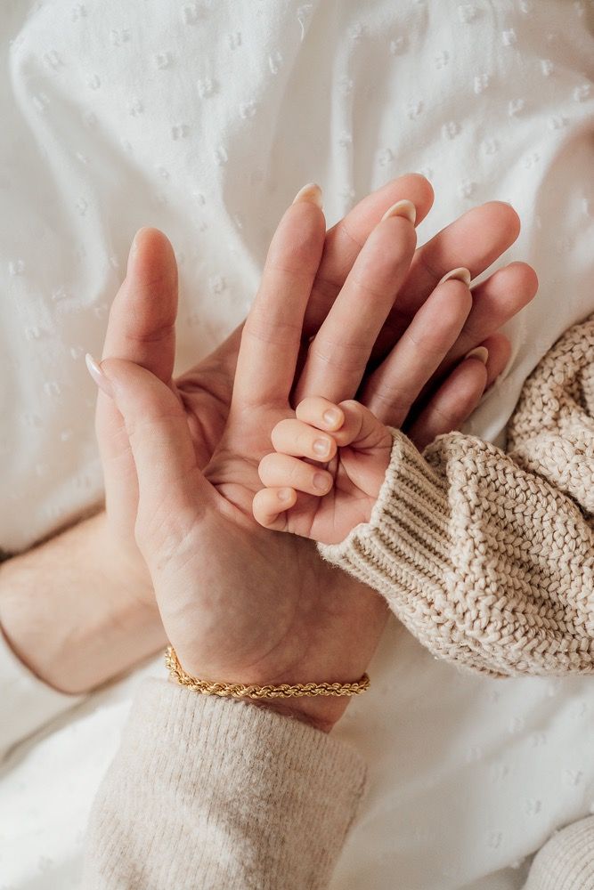 Close up of adult and baby hands holding each other against white bedding, with a knit sweater sleeve and gold bracelet.