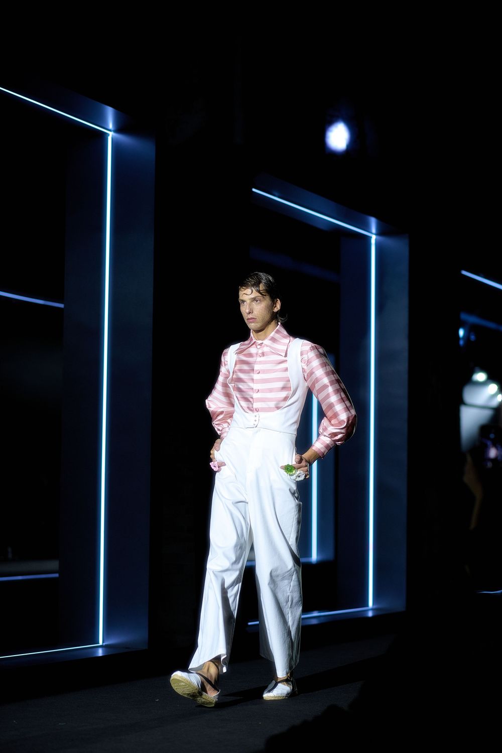 A performer in white pants and striped shirt stands illuminated in a dark space with dramatic blue lighting and shadows.