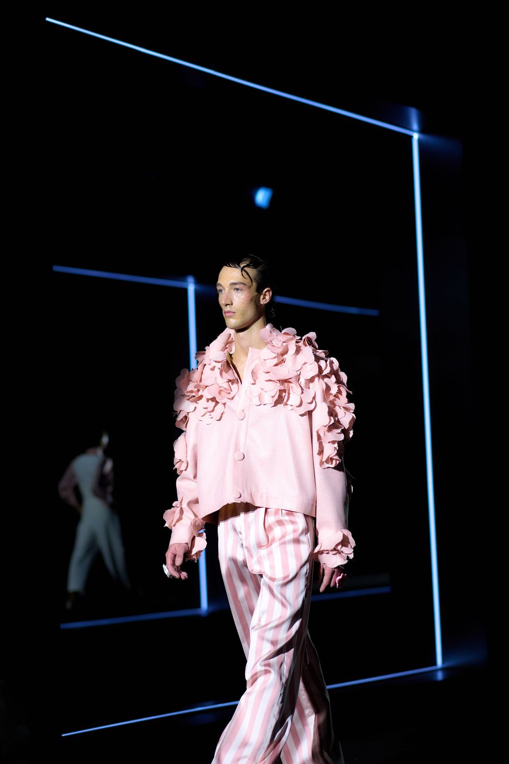 Model walks runway in pink ruffled outfit against dark background with blue neon lighting during fashion show.