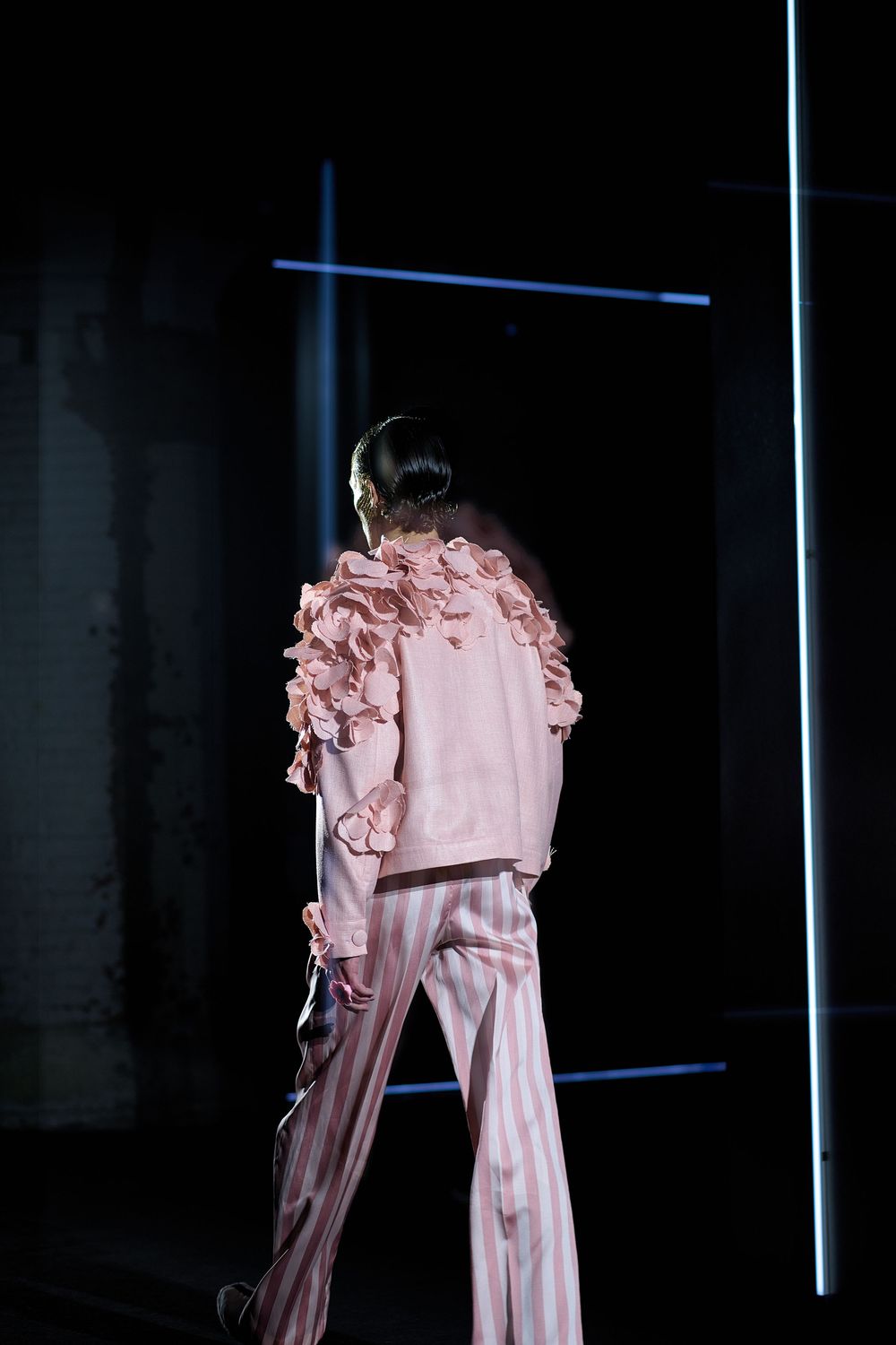 A pink ruffled fashion garment hangs suspended in a dark space with vertical light beams creating dramatic lighting effects.
