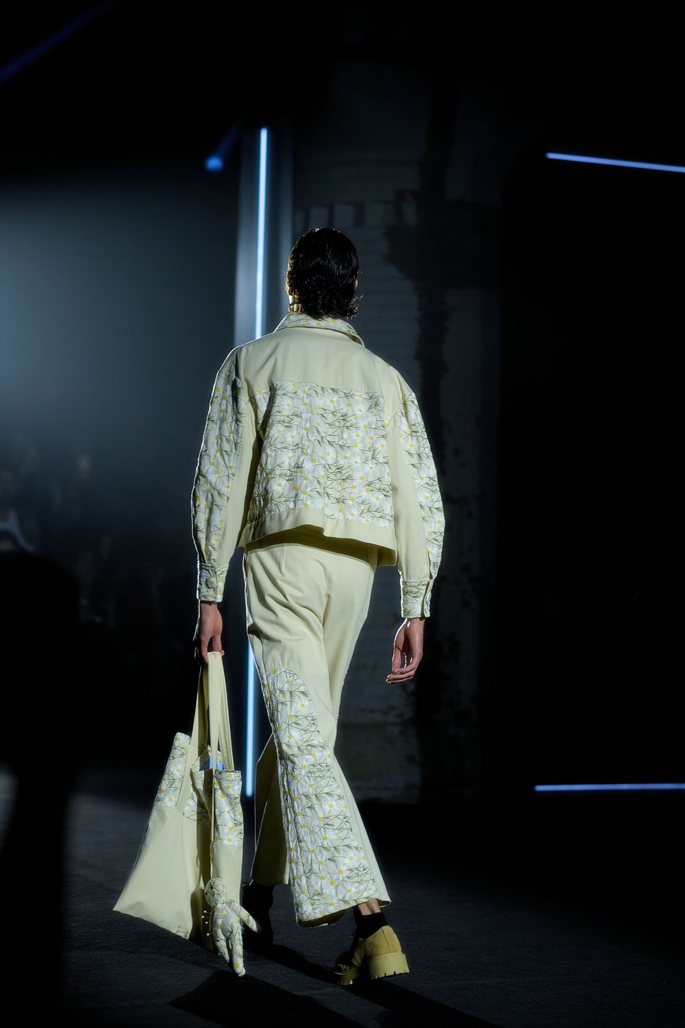 Model wearing a pale yellow embroidered jacket with traditional Asian design details walks on dark runway stage.