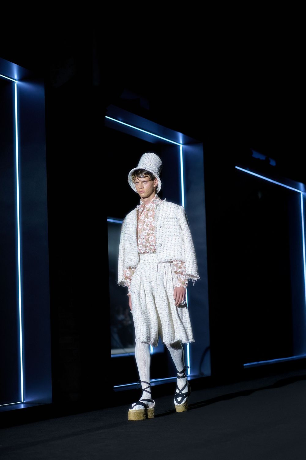 Fashion model in all-white ensemble with platform shoes and bucket hat poses against dark backdrop with blue lighting accents.