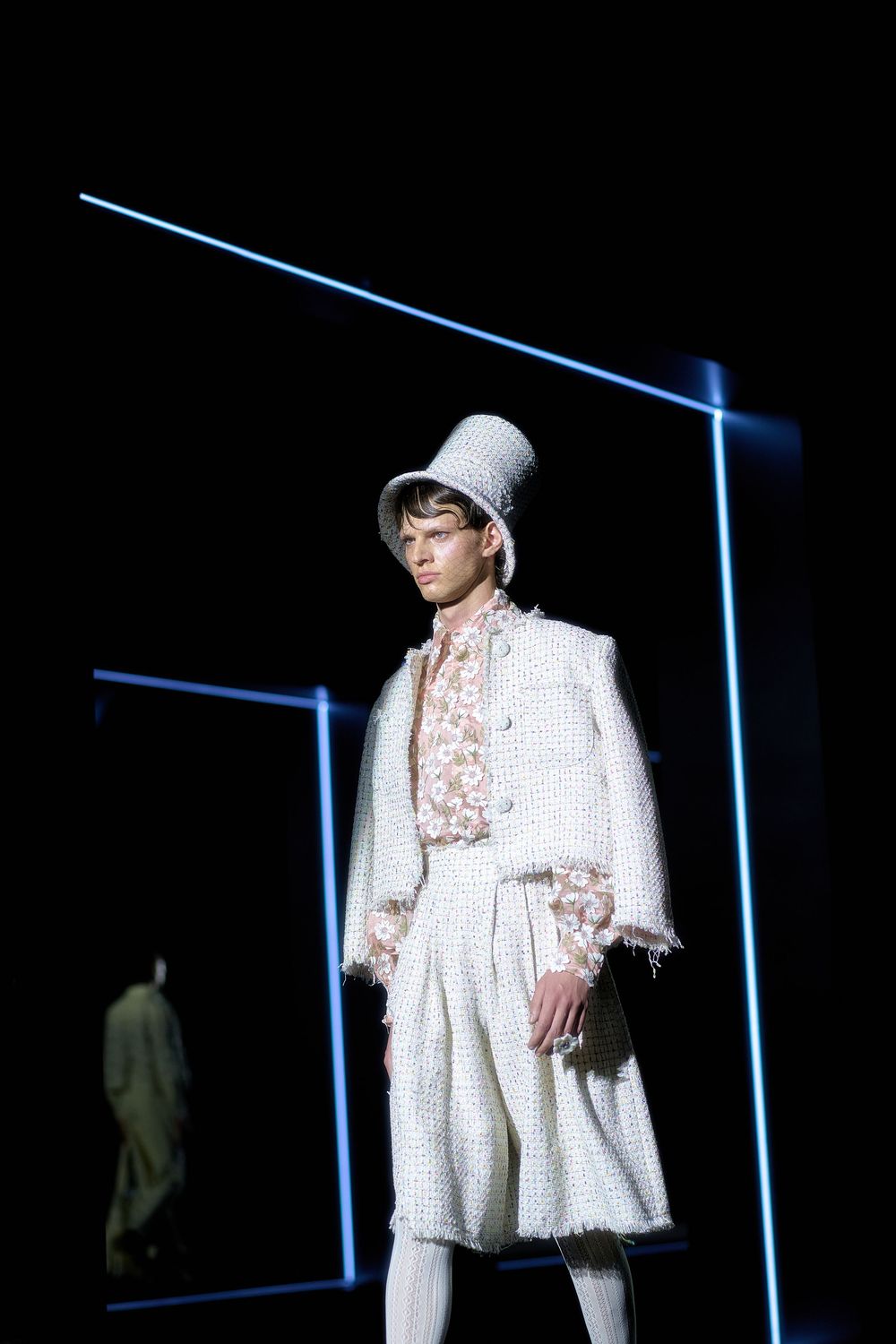 Fashion model walks runway in white oversized dress with geometric patterns and matching baseball cap.