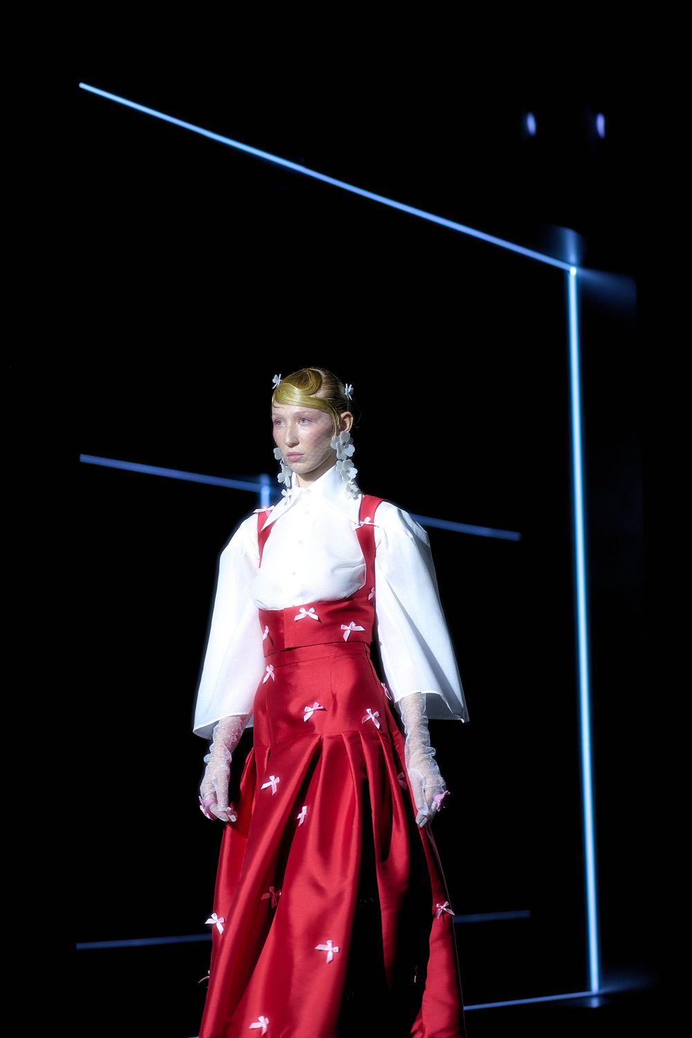 A performer in a red dress and white blouse stands on a dark stage with blue lighting effects in the background.