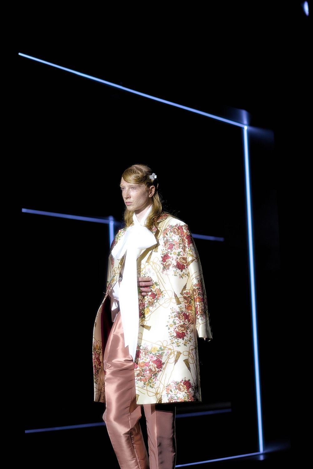 A model walks the runway wearing a floral print coat and pink pants against a dark background with blue neon lines.