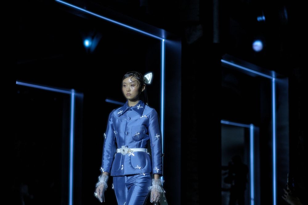 A model walks down a dark runway wearing a sleek blue jumpsuit with white trim during a fashion show.