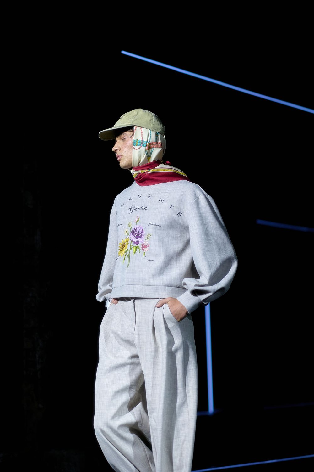 Person wearing a white embroidered sweater, baseball cap and light-colored pants stands against dark background with blue lines.