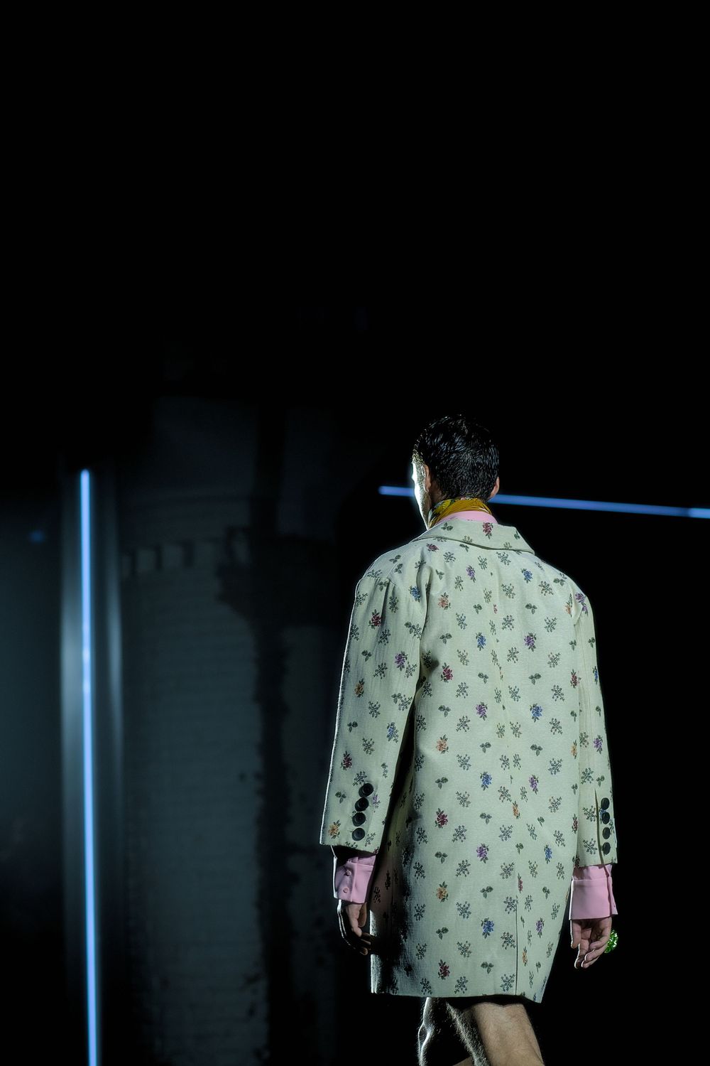 Fashion model walks runway wearing light green patterned coat with pink sleeves in dark moody lighting.