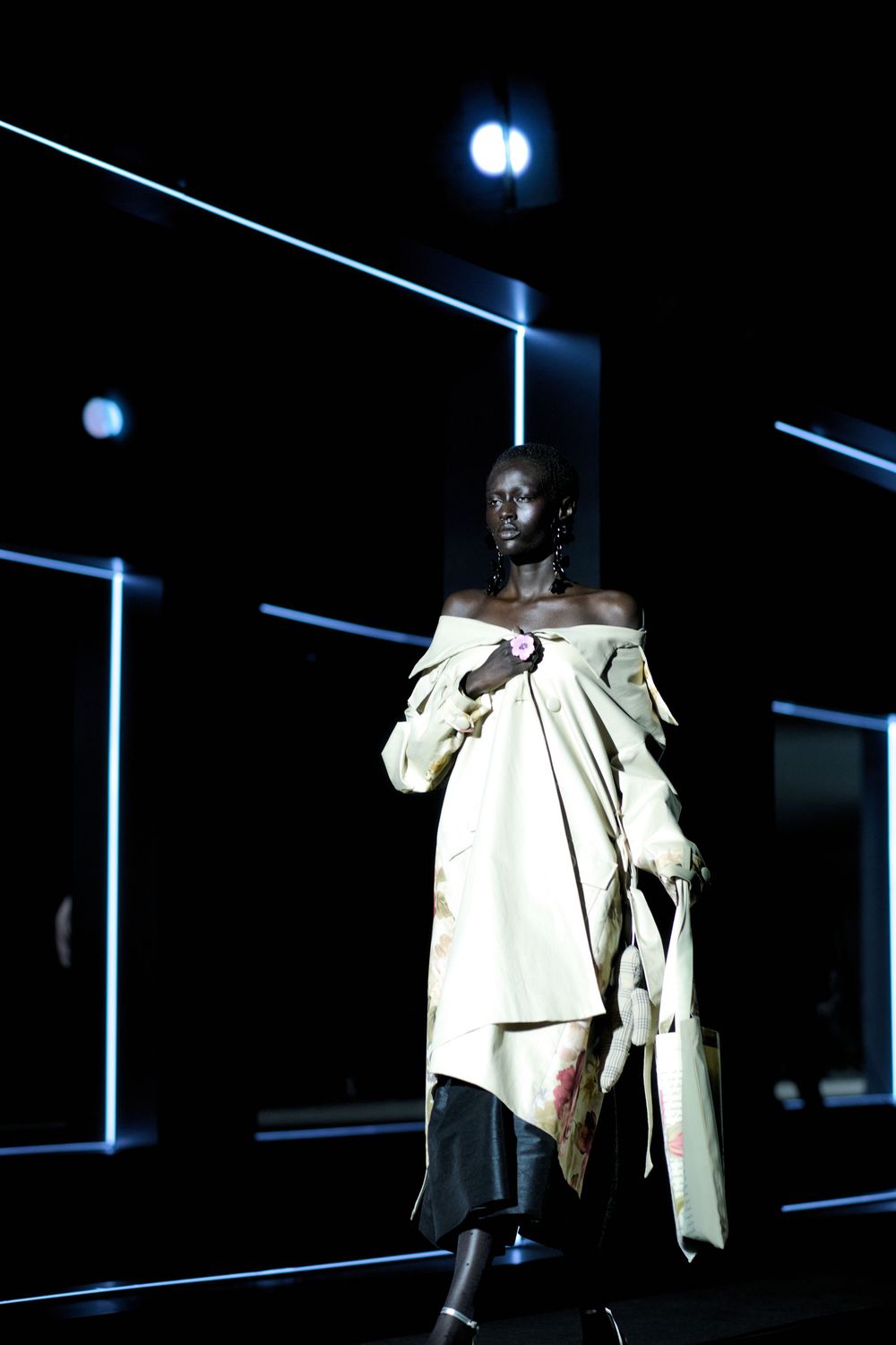 A model walks the runway in an off-white draped garment against a dark background with blue lighting effects.