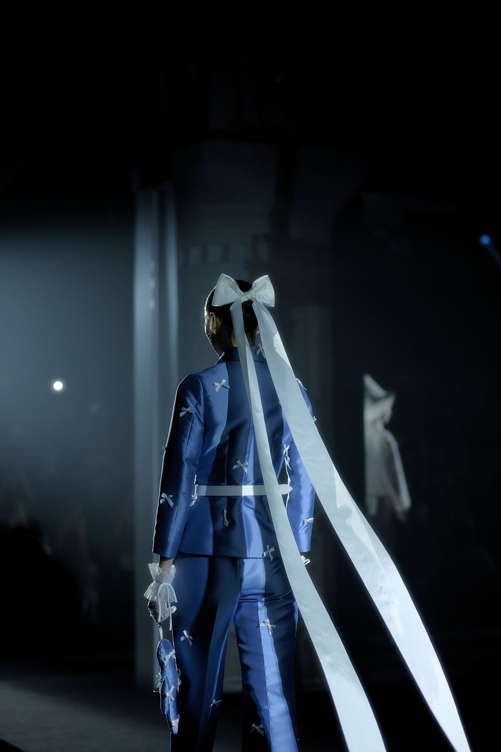 A person in a blue suit jacket stands in a dark room with dramatic lighting and white flowing fabric elements.