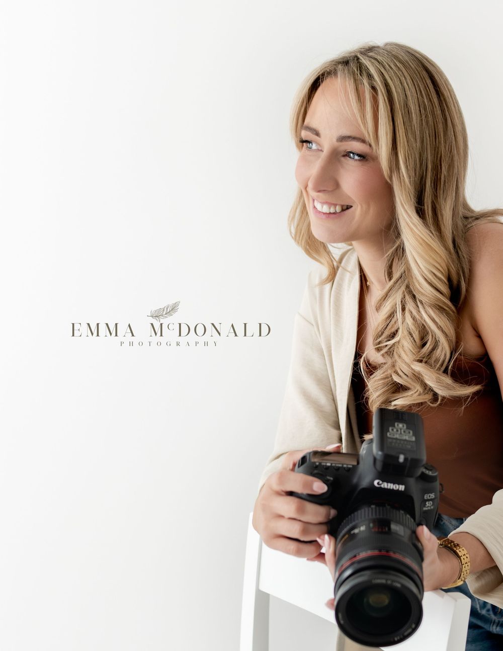 Professional photographer with blonde hair smiles while holding a camera in a bright studio setting.