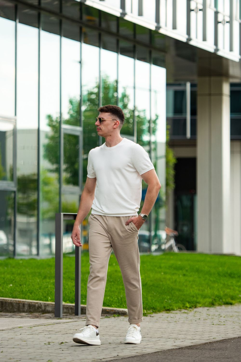 Man in white t-shirt and beige pants poses casually against modern building exterior with glass windows and grass.