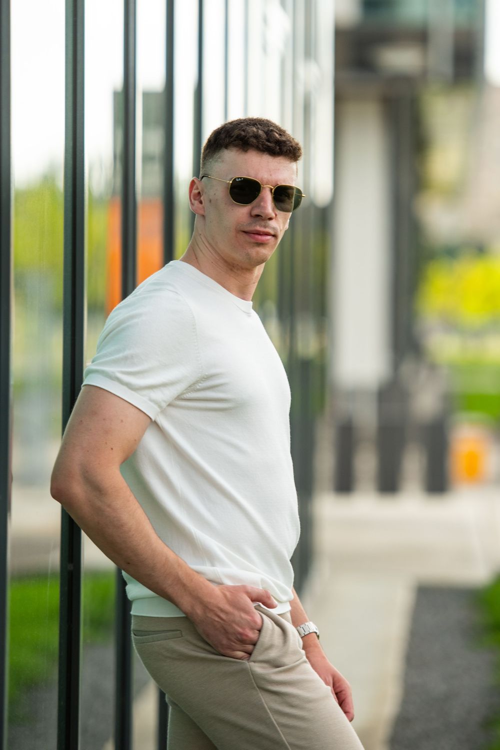 Athletic person in white t-shirt and khaki pants poses confidently against metal railings while wearing sunglasses.