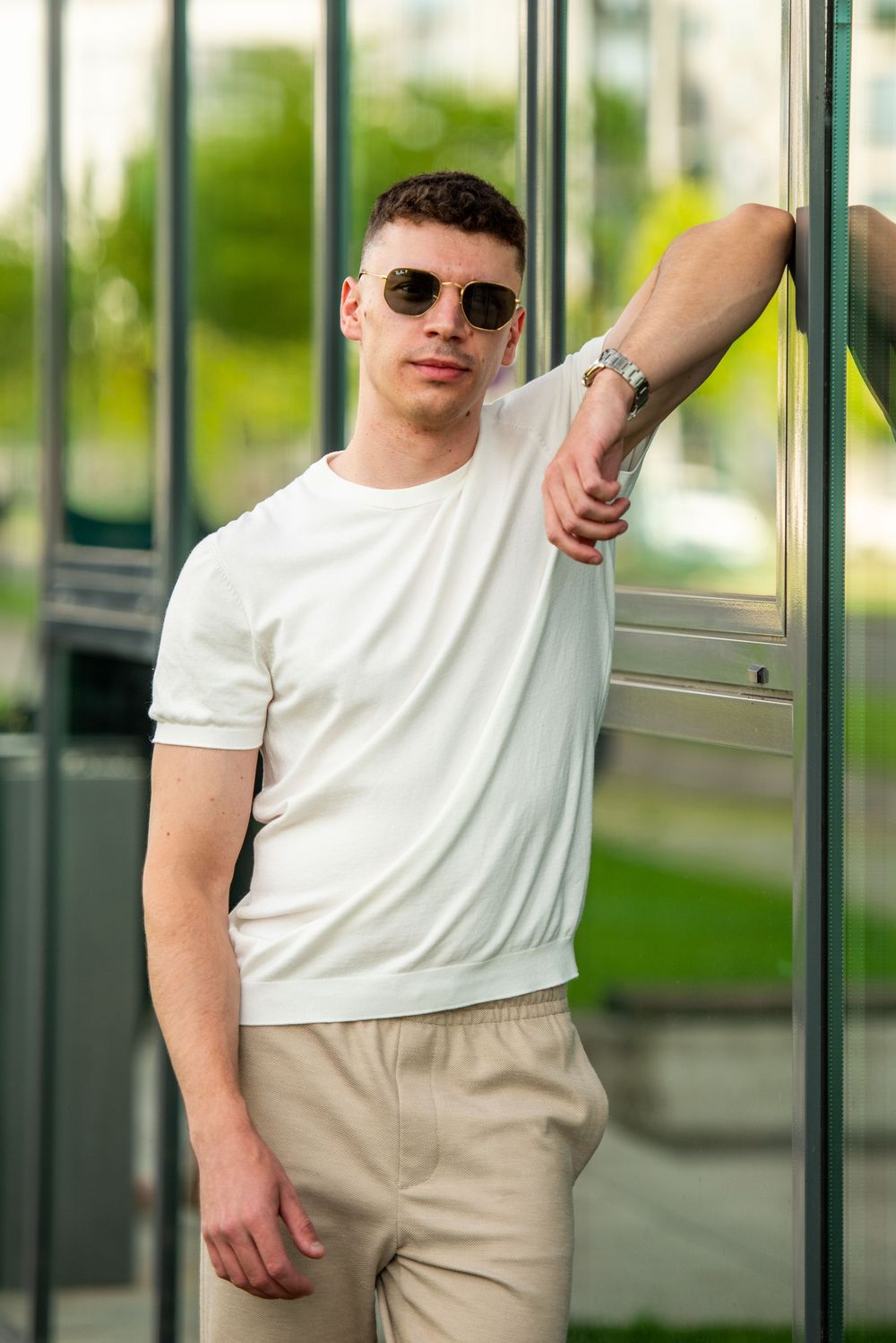 Man in white t-shirt and khaki pants poses casually against glass windows in urban setting.