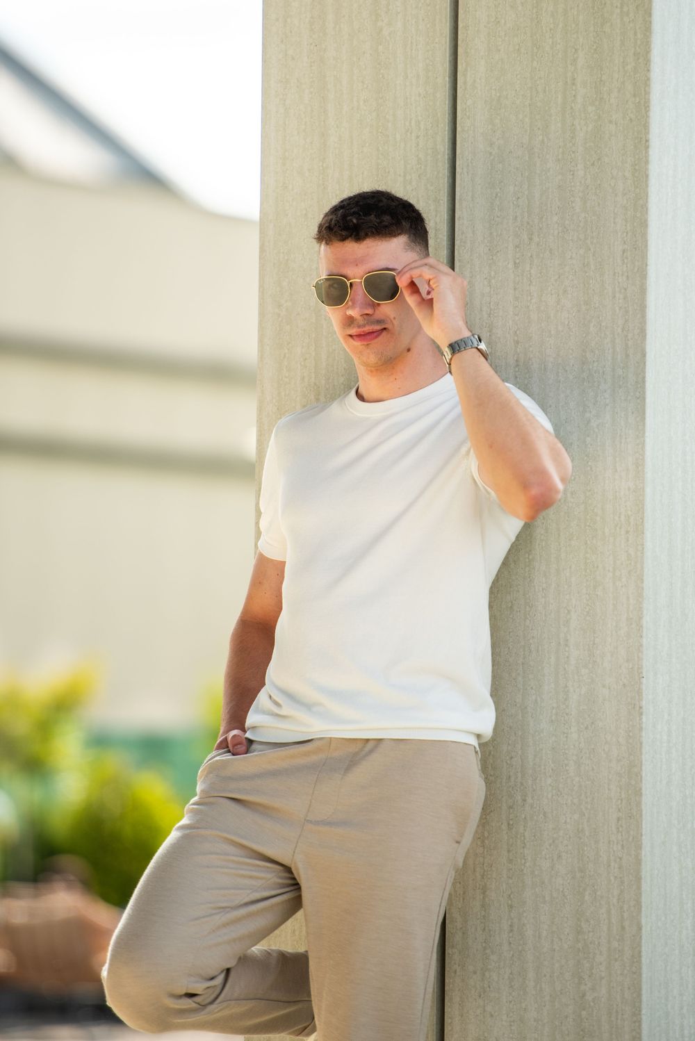 A person in a white t-shirt and khaki pants poses casually against a light-colored column in an outdoor setting.
