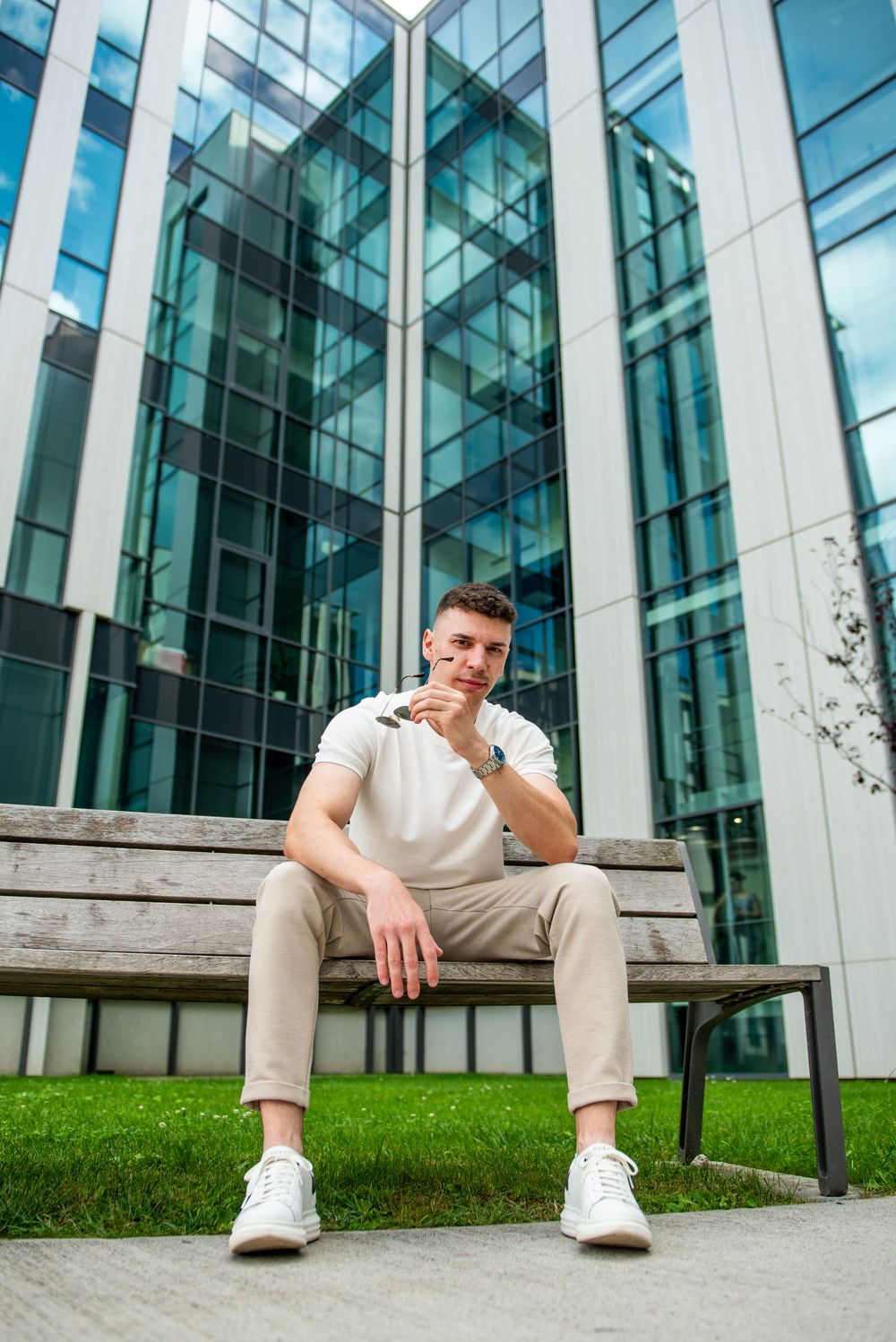 Person in white shirt and beige pants sits on steps outside modern glass office building with green lawn.