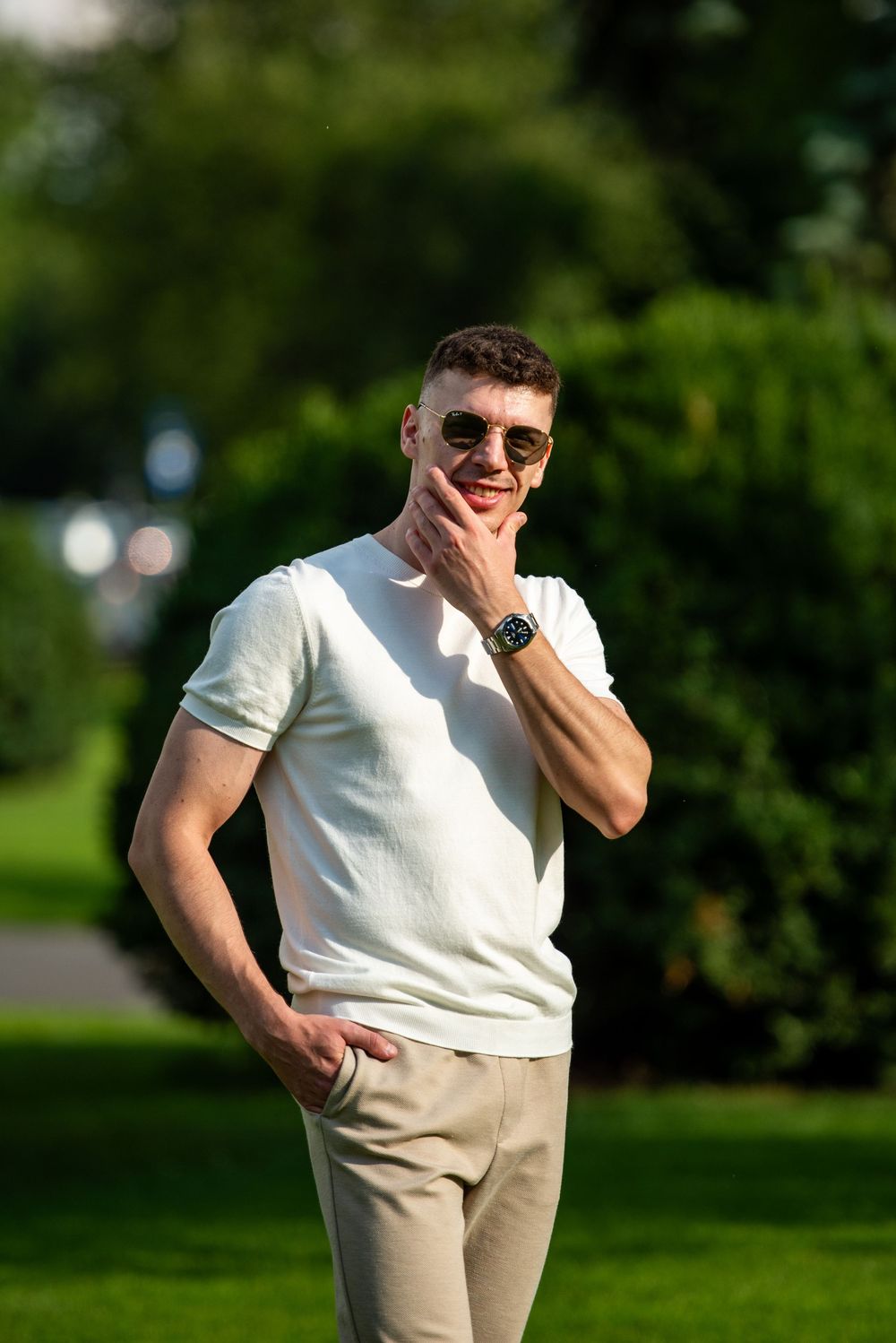 Person in white shirt and khaki pants poses casually outdoors in a sunny park setting.