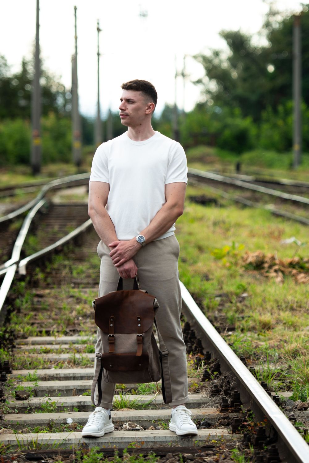 Person in white t-shirt and khaki pants sits contemplatively on train tracks with a brown leather bag nearby.