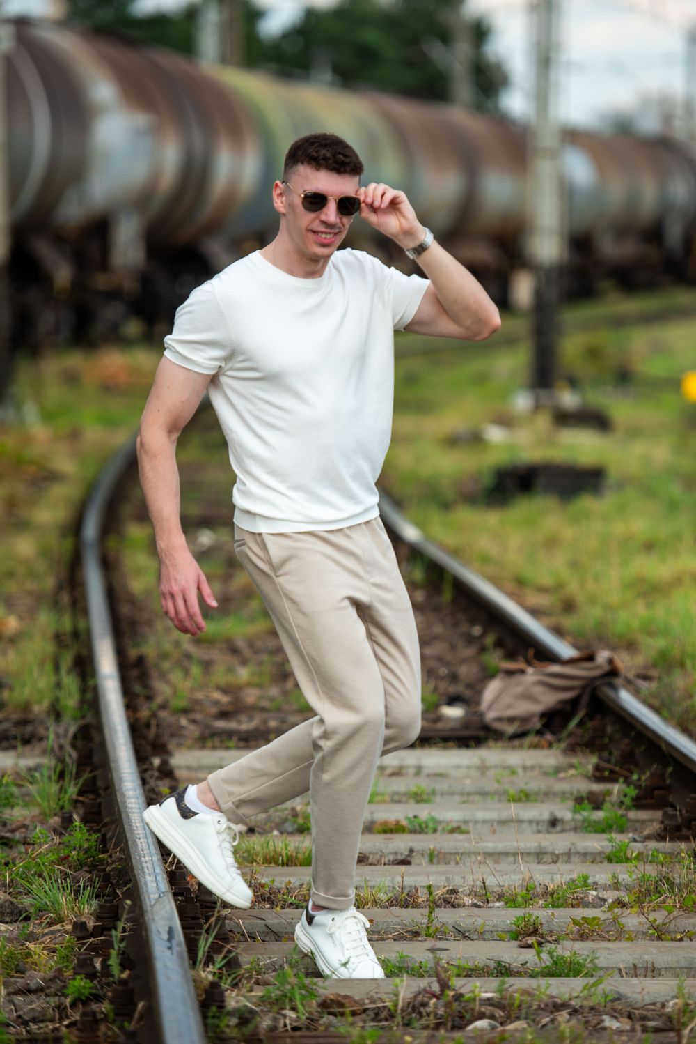 Stylish man in white sneakers and casual attire poses on outdoor steps with railway tracks visible.