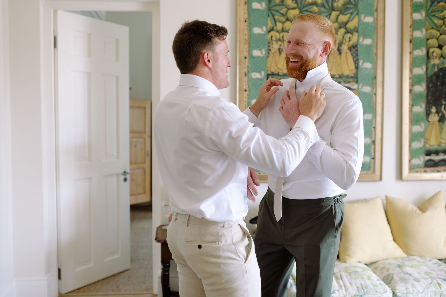 Two people in white dress shirts help each other adjust their bow ties in an elegant room with floral wallpaper.