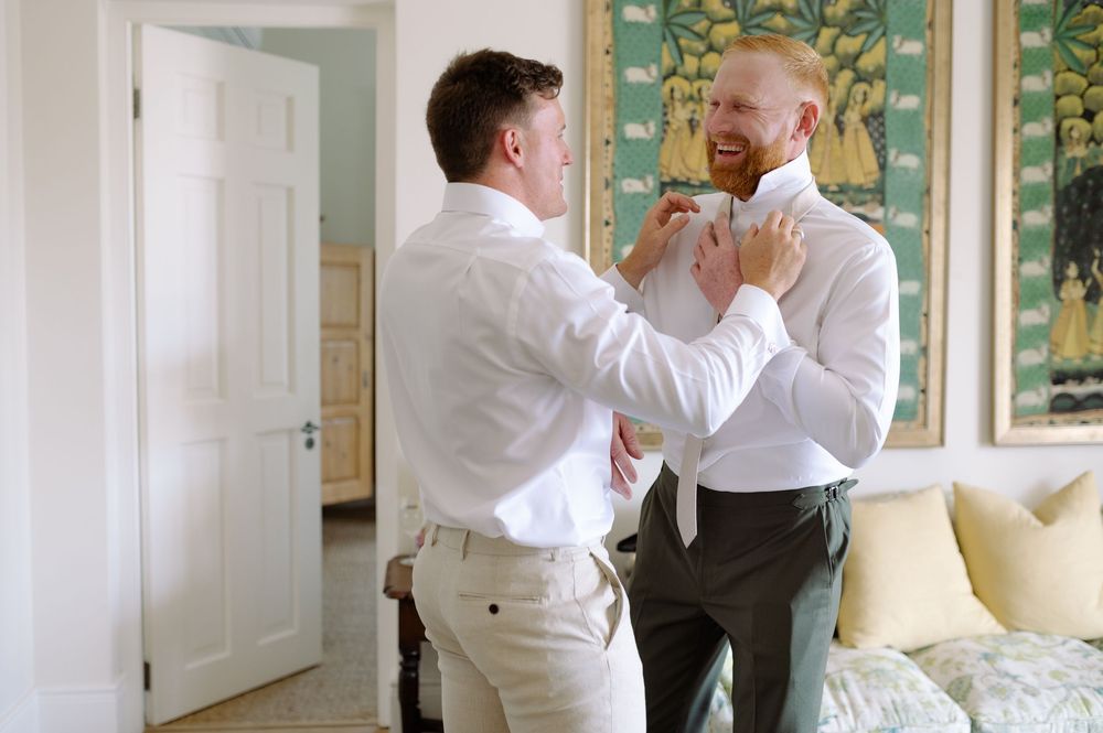 Two people in white dress shirts help each other adjust their bow ties in an elegant room with floral wallpaper.