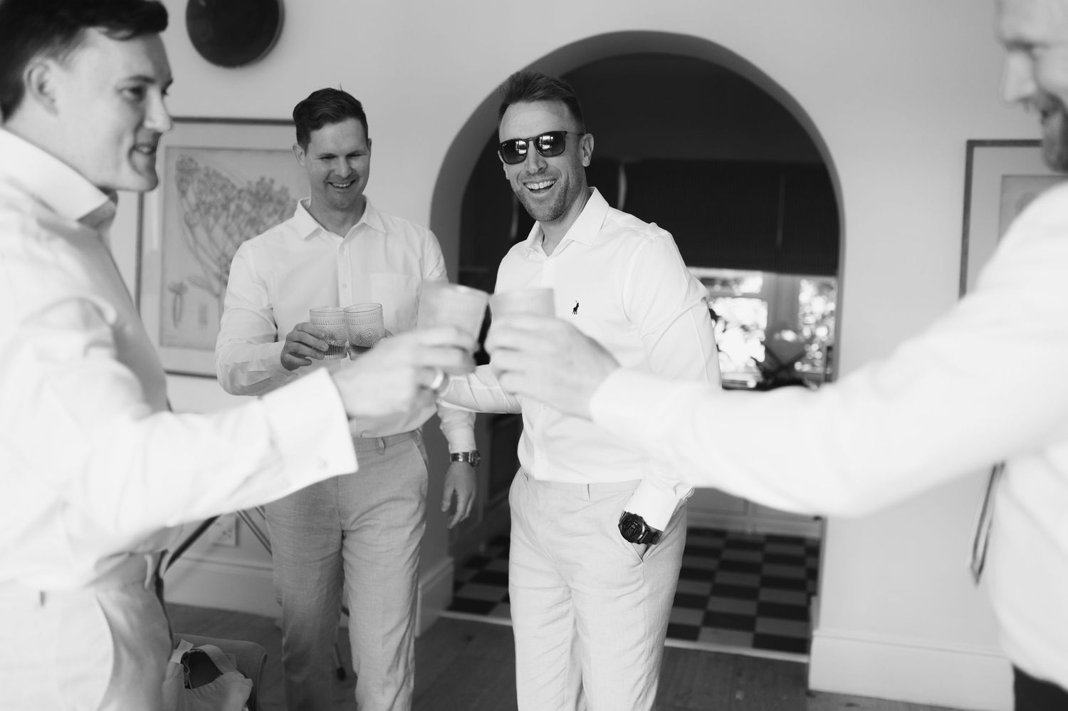 A group of men in white attire and sunglasses gather together in a casual setting with an arched doorway visible behind them.