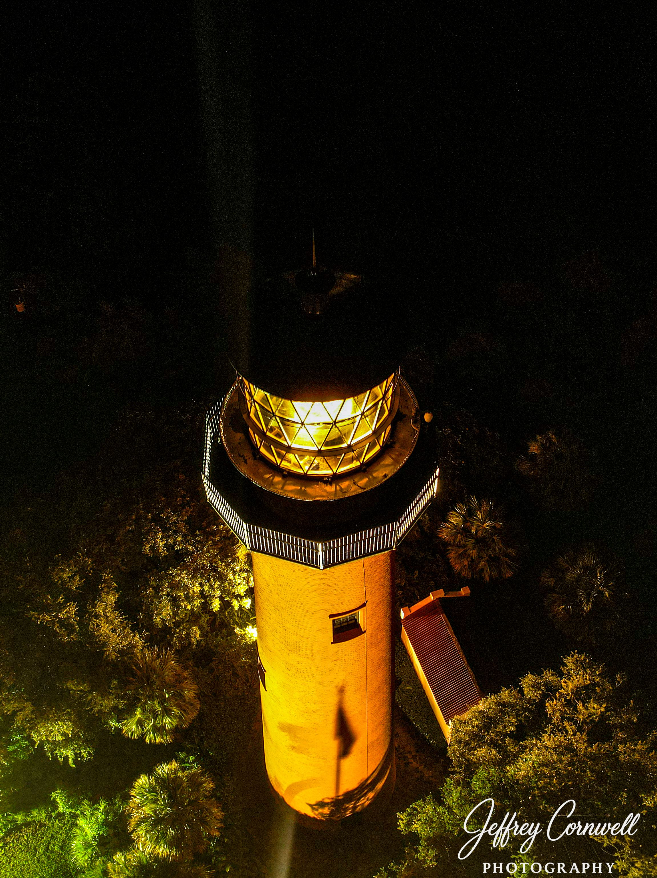 Aerial view of lit lighthouse tower with glowing yellow light chamber and surrounding trees at night.