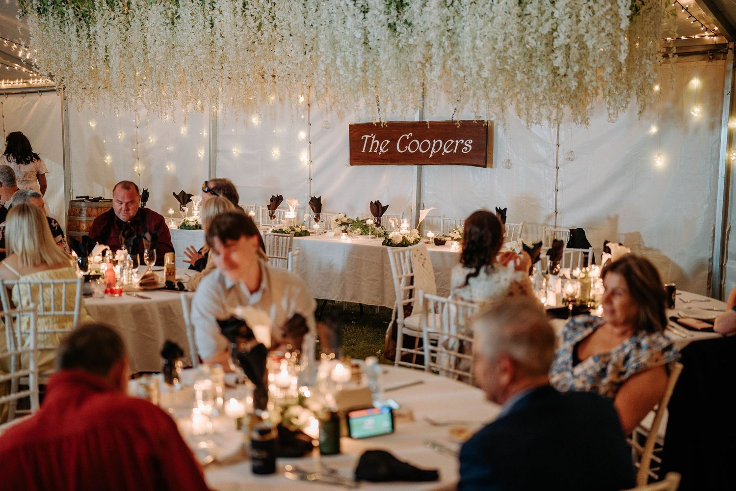 Wedding reception guests seated at candlelit tables under hanging wisteria and string lights at an indoor venue.