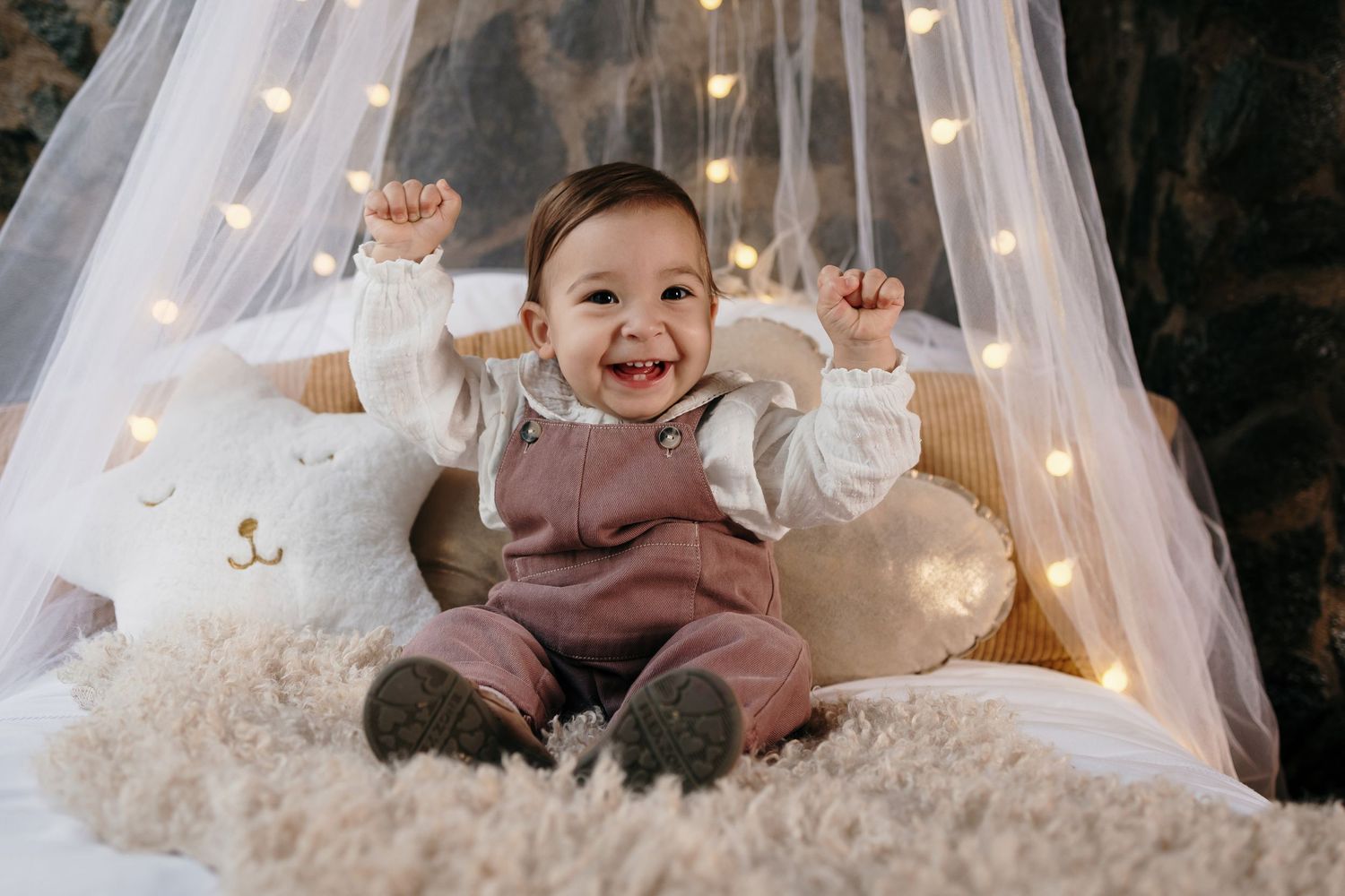 Baby playing on cozy white bedding under sheer canopy with string lights.