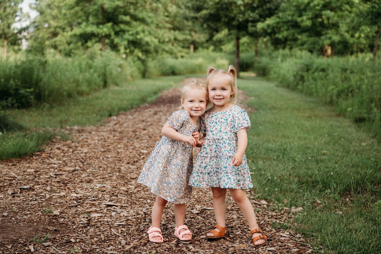 Two infant girls in spring dresses lock arms on a wood chip trail with beautiful greenery.
