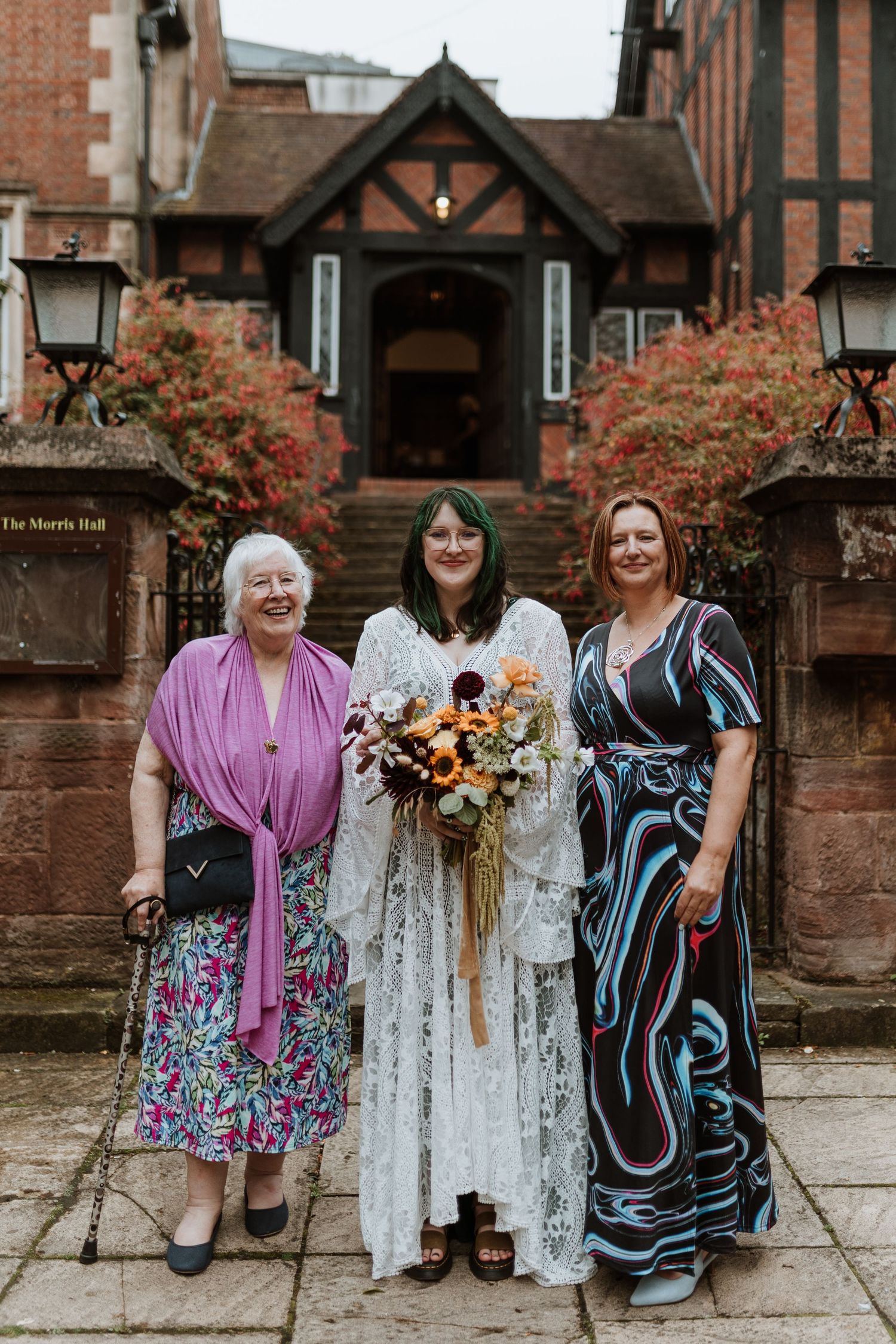 Three women in elegant dresses pose together outside a historic Tudor-style building entrance with autumn flowers.