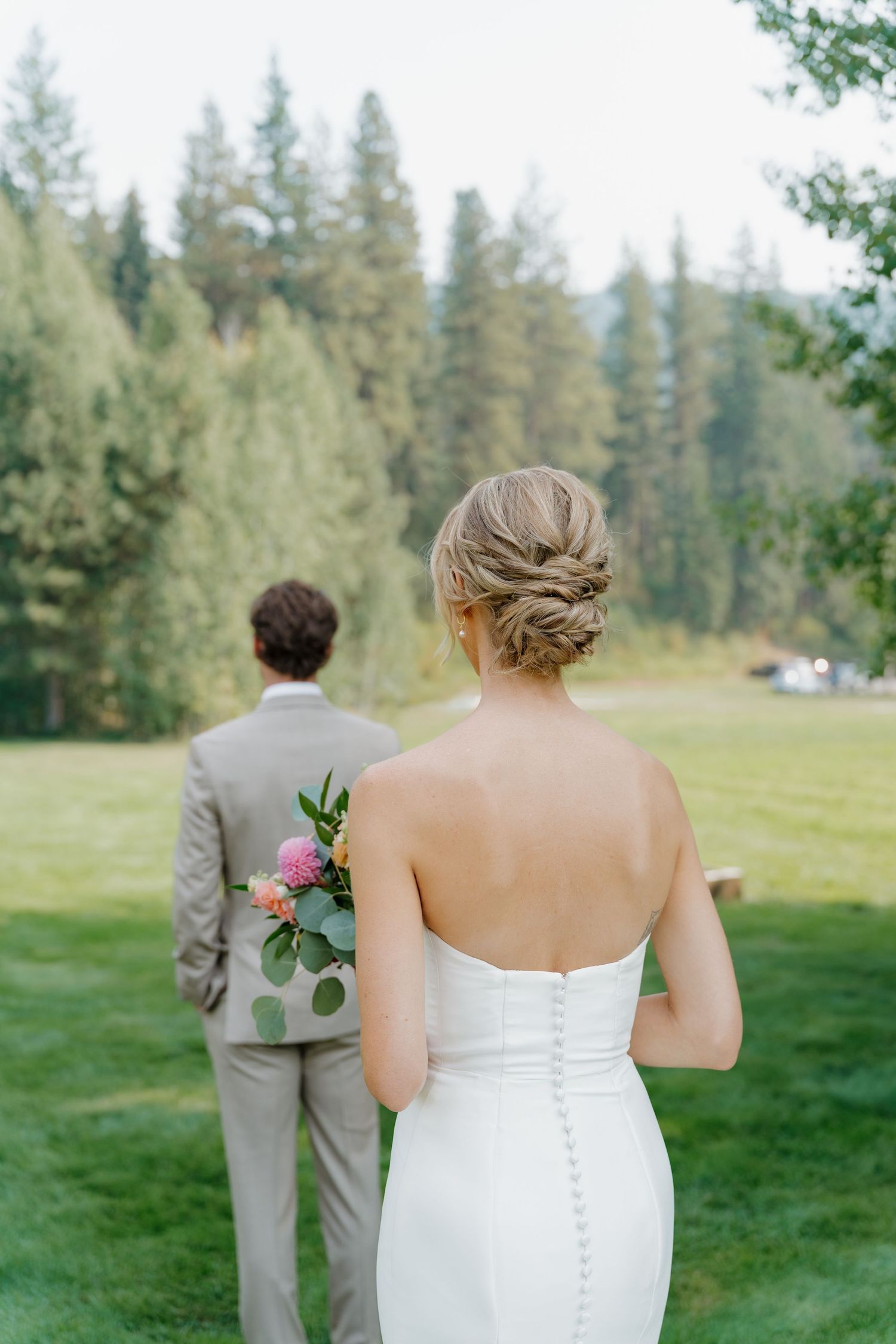 A wedding first look moment unfolds on a grassy lawn with evergreen trees in the background.