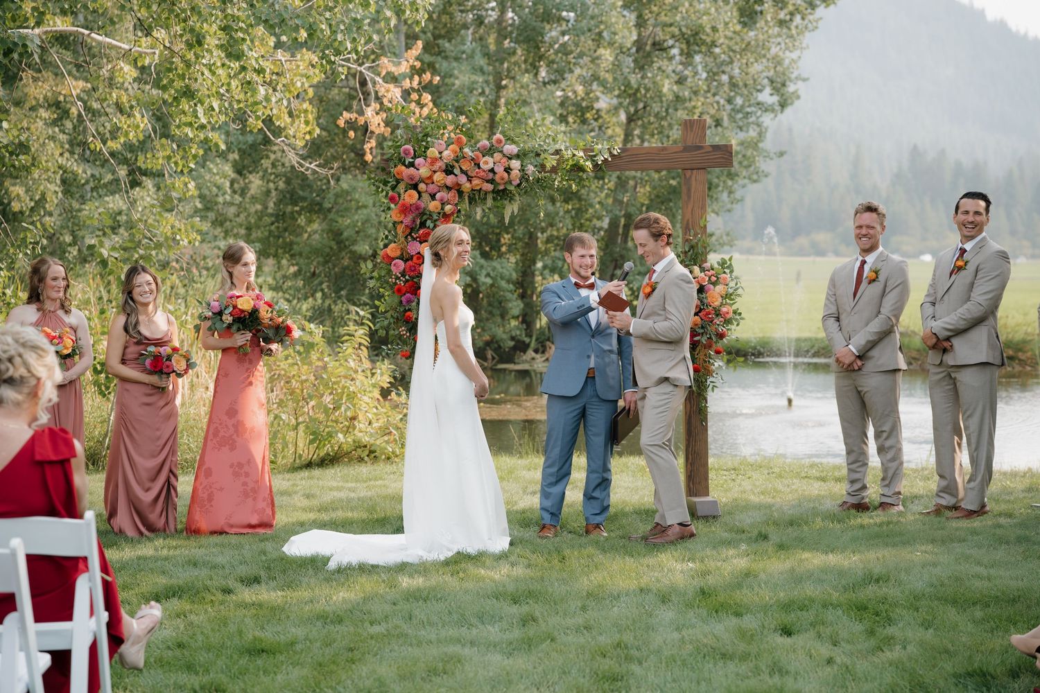 Outdoor wedding ceremony with rustic wooden cross arch and bridal party in pink dresses beside a scenic lake.