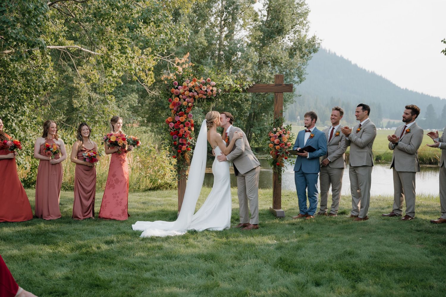 Outdoor wedding ceremony under a floral arch with wedding party in coral and gray attire beside a scenic mountain view.