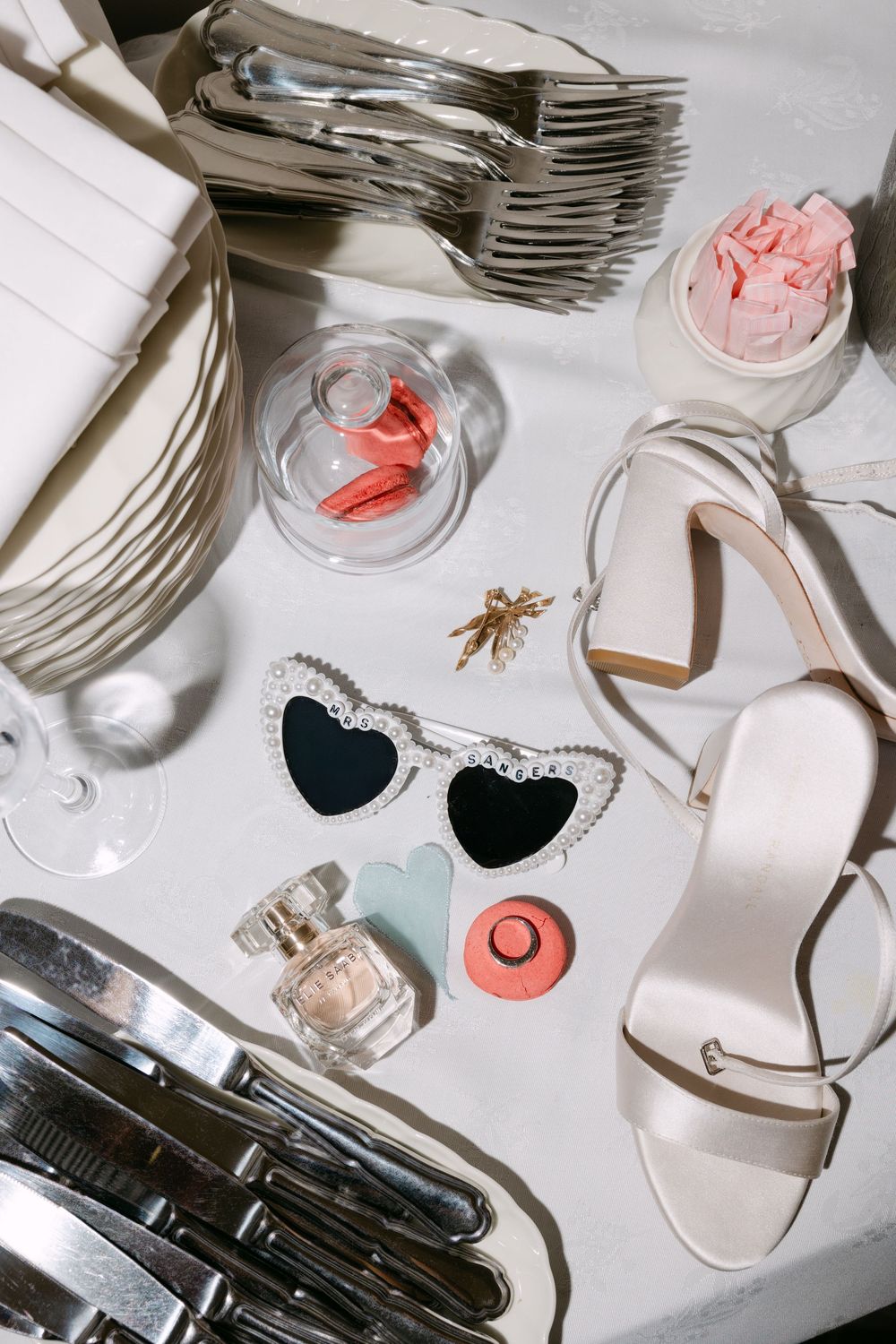 Wedding accessories including heart sunglasses, perfume, shoes, and silverware arranged on white surface.