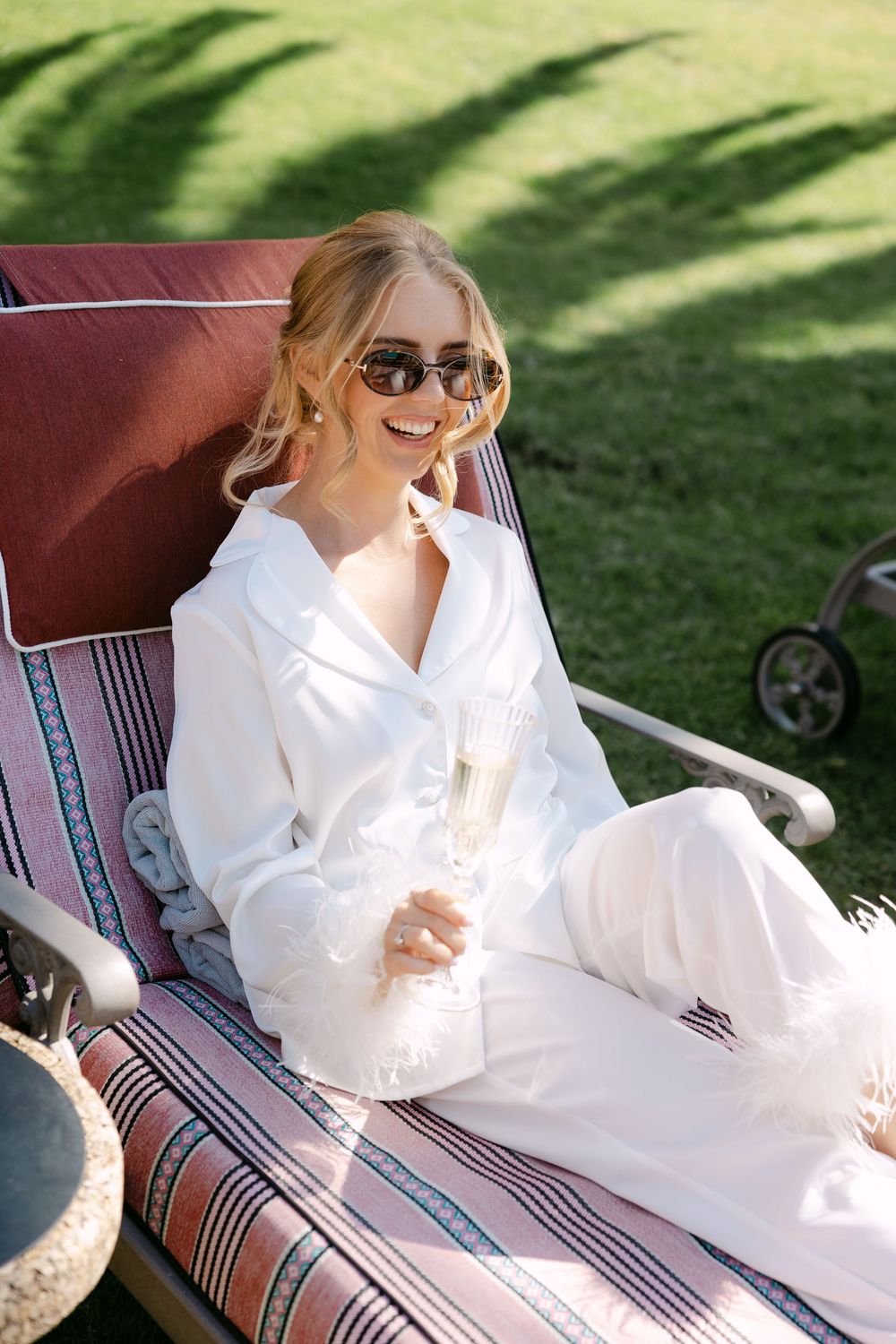 A person in a flowing white dress lounges on a striped outdoor chaise lounge chair in dappled sunlight.