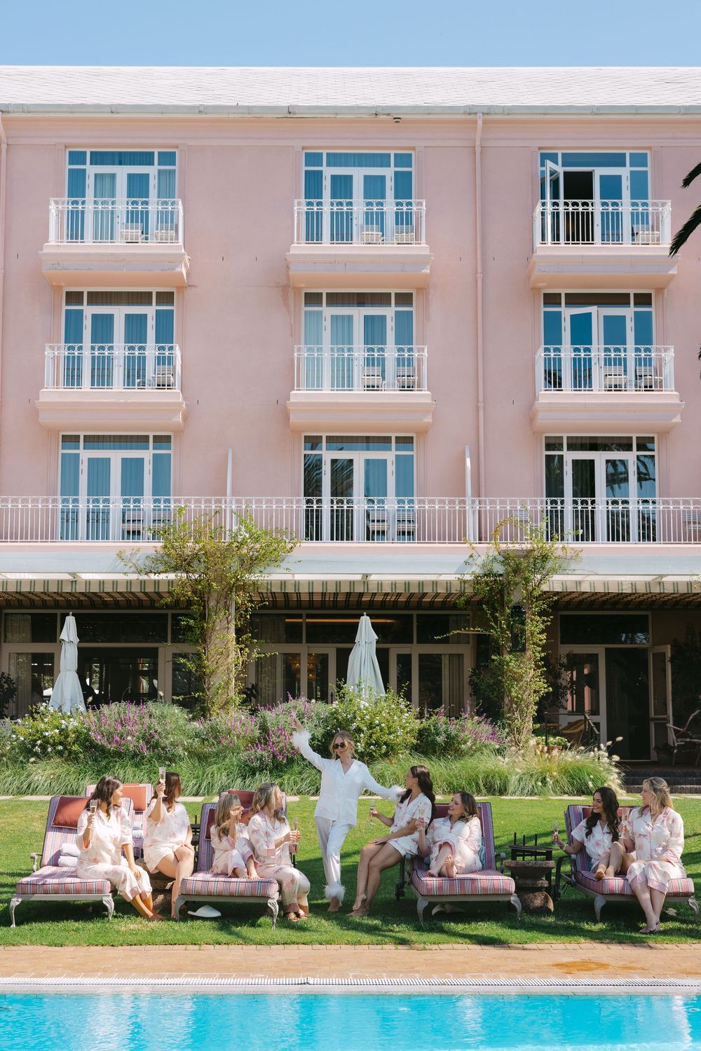 A group gathers on the lawn by a pool outside a pink hotel with white balconies and tropical landscaping.