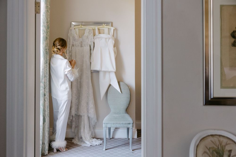 A white wedding dress and suit hang in a bright dressing room with a light blue chair and framed artwork on the wall.