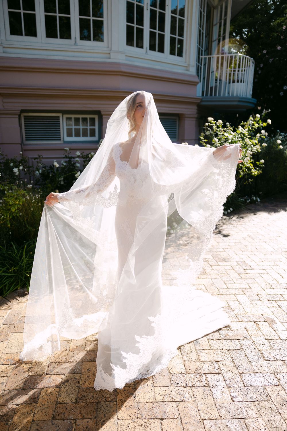 Ethereal bridal portrait with flowing veil against pink building and brick pathway in sunlight.