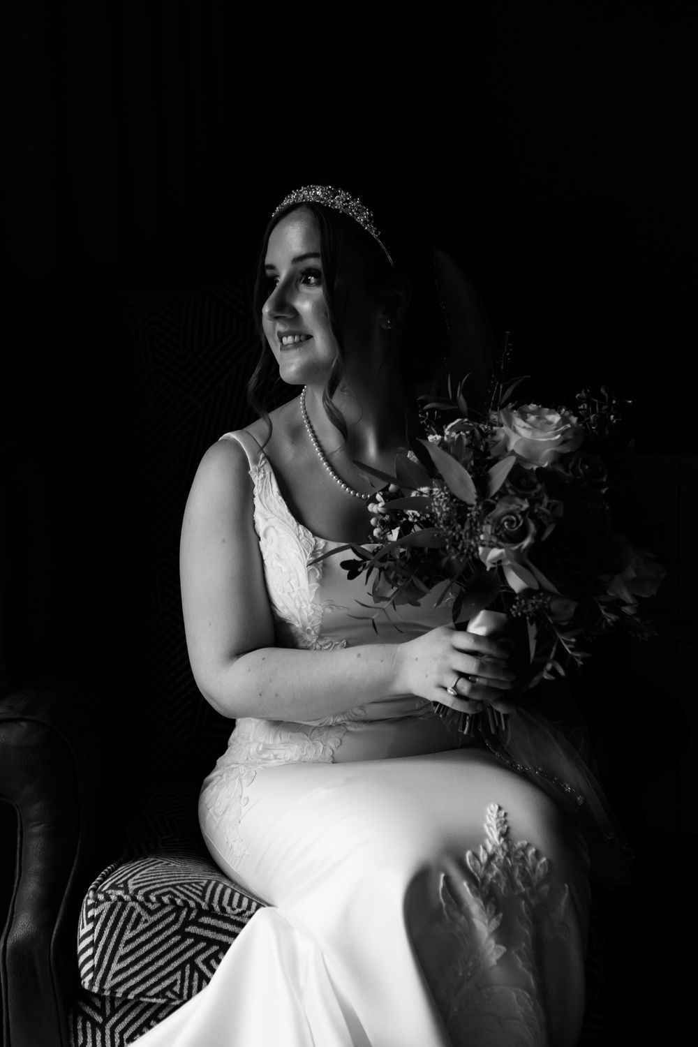 A dramatic black and white photograph of a bride in a white dress sitting with a bouquet against a dark background.
