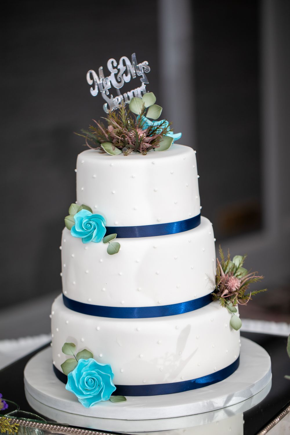 Three-tiered white wedding cake decorated with blue roses, navy ribbons, and a custom cake topper.